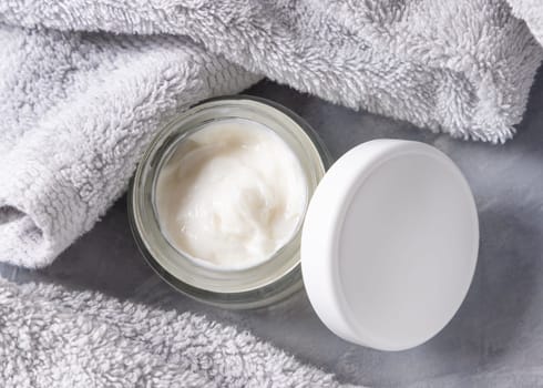 White opened cream jar with blank lid near light grey towel in bathroom top view, brand packaging mockup. Natural organic cosmetics for daily facecare and spa procedures, copy space