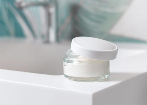 Opened cream jar with white blank lid on washbasin with faucet in green tropical bathroom close up, packaging mockup. Lifestile scene with ubranded natural facecare product