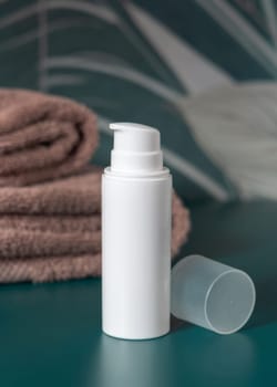 White cream one pump bottle near brown folded towels in dark green tropical bathroom close up, packaging mockup. Natural plant unbranded cosmetic container, copy space. Spa and wellness concept