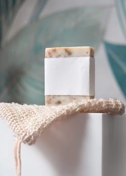 Beige handmade soap bar with blank label on sisal soap saver bag on washbasin in green tropical bathroom close up, mockup, copy space. Natural herbal unbranded product for daily hygiene