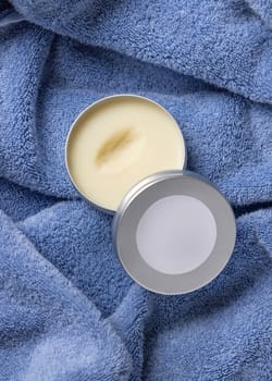 Opened body butter jar with blank label on blue bath towel in bathroom top view. Natural herbal homemade cosmetics for body care, daily beauty routine and spa procedures