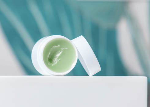 Opened white blank lip balm jar with a lid on washbasin border in green tropical bathroom close up, packaging mockup. Lifestile scene with ubranded natural lipcare product