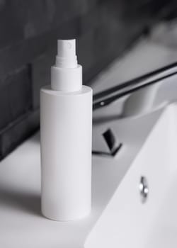 White spray bottle near modern faucet on basin in dark grey masculine bathroom close up, packaging mockup. Natural unbranded cosmetics with copy space. Spa and wellness concept