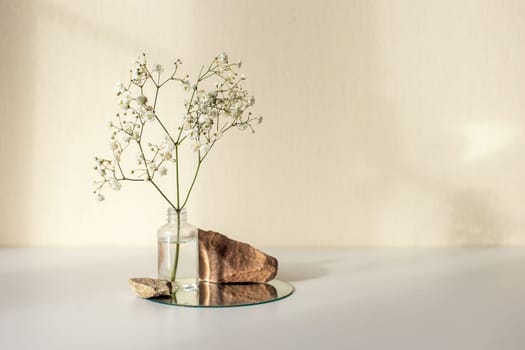One branch of a white gypsophila flower standing on a little bottle and on a round mirror with stone on a beige background, copy space, abstract design concept. Mock up