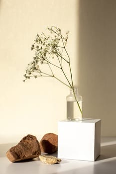 Simple geometric minimalism still life with a branch of a gypsophila flower standing on a white paper cube and three stones on a beige background, copy space, abstract design concept.
