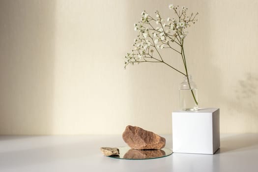One branch of a gypsophila flower standing on a white paper cube, near three stones on a round mirror, on a beige background, copy space, abstract design concept. Mock up