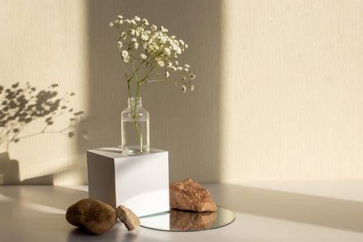 A branch of a gypsophila flower standing on a bottle on a white paper cube, near three stones on a round mirror, on a beige background, copy space, abstract design concept.