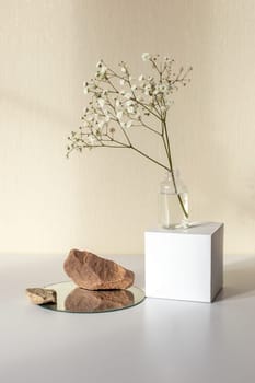 One branch of a gypsophila flower standing on a white paper cube, near three stones on a round mirror, on a beige background. Mock up. Vertical