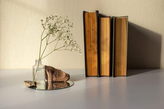 Three old books stand next to a branch of a white gypsophila flower standing in a small bottle and on a round mirror with stones, on a beige background, copy space, abstract design concept.