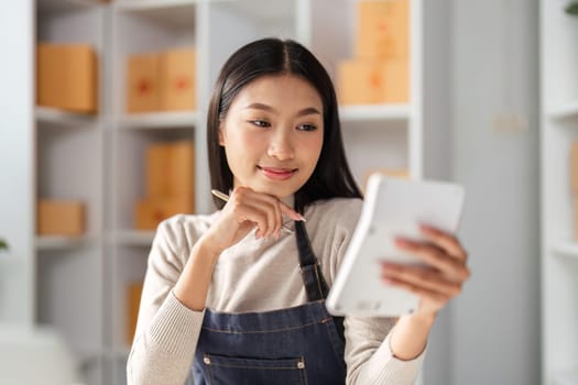 A young female entrepreneur reviews her business on a tablet, showcasing focus and dedication in a modern workspace filled with boxes.