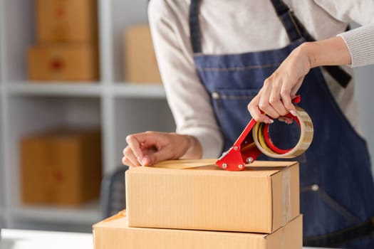 Close-up of a young woman's hands sealing a shipping box with tape in a bright and organized workspace filled with boxes.