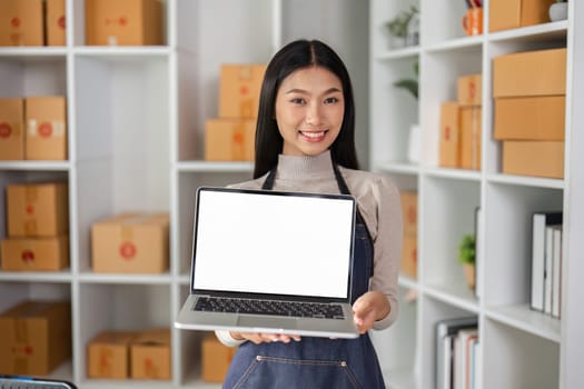A smiling young female entrepreneur presents a laptop in a modern workspace, highlighting her business and creativity.