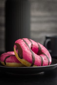 Sweet pink donut with chocolate strips on plate on a black table.