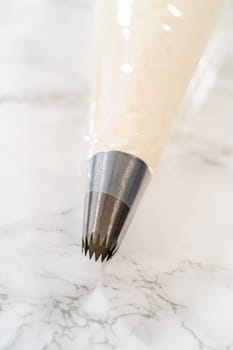 Poised on a marble surface, the piping bag's star nozzle is filled with creamy eggnog buttercream, shining under a bright light.