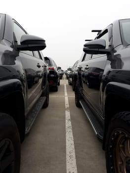 Vertical photo between two black SUVs in street parking lot for new cars. Car market on street with many parked cars for sale. Abstract photograph of row of cars on street