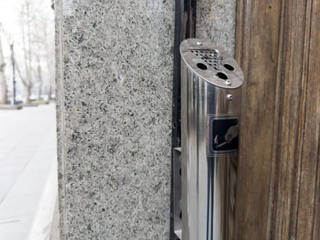 Outdoor ashtray is attached to building for convenient smoking of cigarettes. Ashtray and trash can in smoking area. Bad habit of smoking cigarettes. Steel trash can post at entrance to building