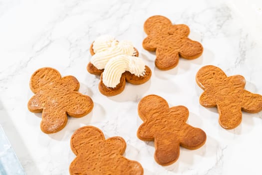 Gingerbread cookies await their second halves on a marble surface, each meticulously piped with buttercream to craft delightful sandwich treats. The precision of the piping adds a festive touch.