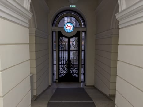 Corridor to exit door in Georgian building with symbol of country above large black doors with glass windows. Georgian flag at exit of building through arch and large doors