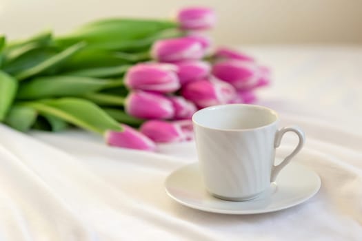A white cup with a saucer and a bouquet of beautiful pink tulips lies on a white tablecloth. close up. copy space