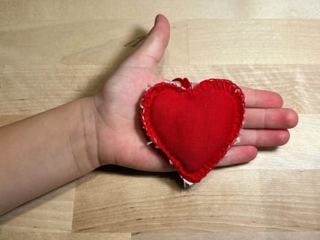 Heart sewn from red fabric in the hand of a small child on a wooden background. Love
