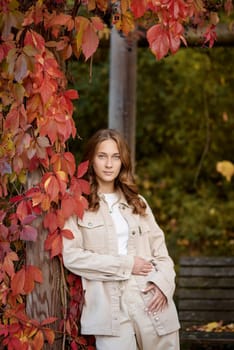 A radiant young woman in a casual beige ensemble leans gracefully against a rustic wooden pergola adorned with vibrant red autumn leaves. The warm, golden tones of fall foliage create a natural backdrop, highlighting her joyful smile and the inviting atmosphere of the garden setting. This image captures the essence of autumn lifestyle, combining modern casual fashion with the organic textures of seasonal nature. Perfect for promoting outdoor living, fashion blogs, travel content, or autumn-themed marketing campaigns, the scene evokes feelings of relaxation, connection with nature, and the comforting charm of the fall season. A young woman with wavy light brown hair and a cheerful smile is leaning against a wooden pillar wrapped in red autumn leaves. She is dressed in a beige casual outfit, including a stylish denim jacket and matching pants, with a white top underneath. The background features a scenic garden pergola with wooden beams covered in lush green and red foliage, creating a natural and cozy atmosphere. The warm lighting and soft depth of field enhance the autumn aesthetic, making this image perfect for seasonal fashion, lifestyle blogs, outdoor portraiture, and autumn-themed marketing campaigns.