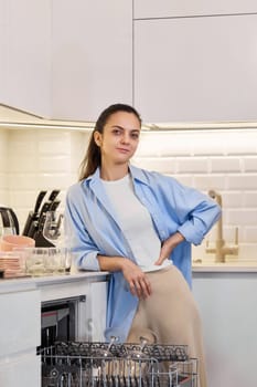 Happy woman in blue shirt standing near open dishwasher in a white modern kitchen