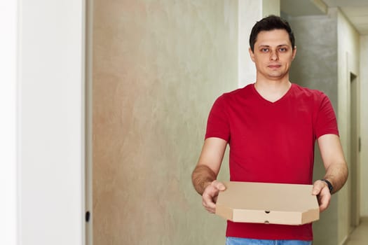 handsome delivery man in red t-shirt giving food order, holding pizza box standing at doorway, copy space