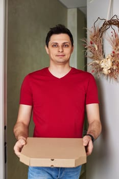 positive delivery man in red t-shirt giving food order, holding pizza box standing at doorway
