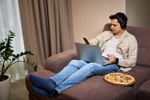 Young man sitting on sofa and eating pizza while working on laptop