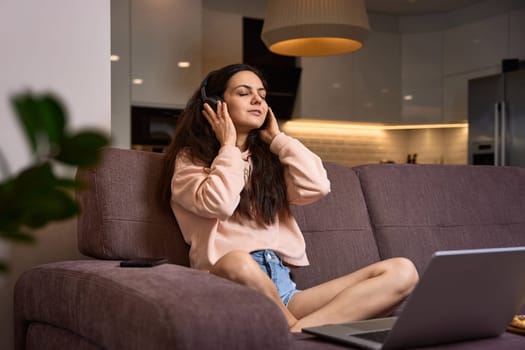 happy Caucasian woman listening to music with headphones and using laptop on couch at home