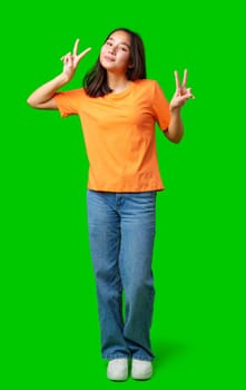 A young woman is standing confidently with both hands forming peace signs. She wears an orange shirt and blue jeans, showcasing a joyful expression.