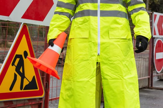 A worker wearing a bright yellow high-visibility raincoat and safety boots places an orange traffic cone on a wet asphalt road.