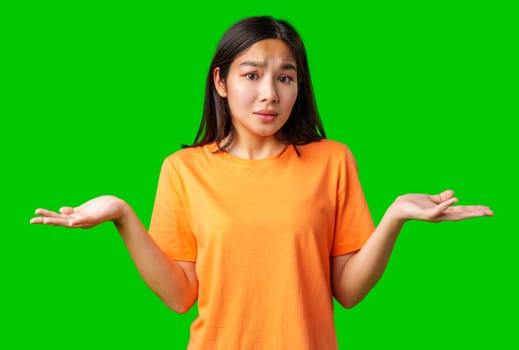 A young woman stands against a green background, wearing an orange shirt, raising her hands in a gesture of confusion or uncertainty, as if contemplating her next move or decision.