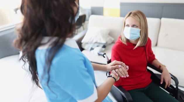 Woman in wheelchair in protective medical mask gives hands to doctor. Coronavirus and disability concept