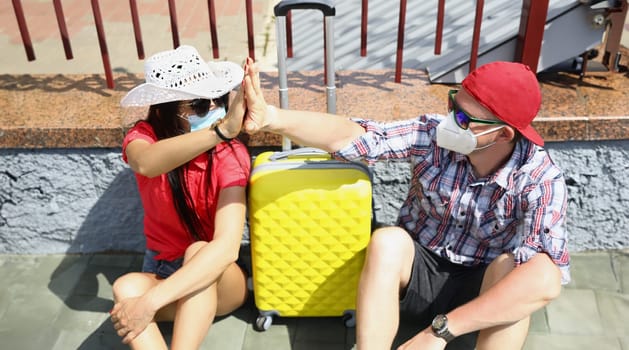 Man and a woman in medical protective masks sit at train station with suitcase and give bad friend five. Traveling around the world in the coronavirus pandemic concept