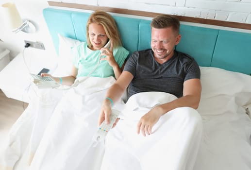 Man and woman are lying on bed. Woman is talking on phone, man switches buttons on remote control. Features of the hotel industry concept