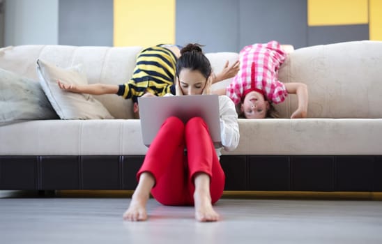 Tired woman with laptop sits on floor with closed palms ears behind on couch children indulge. Remote work in coronavirus pandemic concept