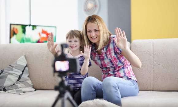 Joyful mother and daughter hold their hand in greeting to video camera screen. Family video blogging concept