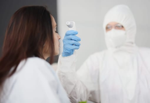 Doctor in protective suit and gloves measures temperature of person. Temperature measurement with non-contact thermometer in the coronavirus pandemic concept