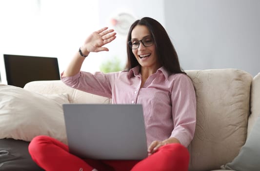 Smiling woman wearing glasses waving at laptop monitor while sitting on sofa. Online broadcast in social networks in quarantine concept