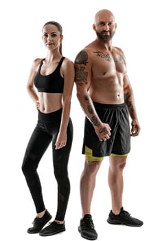 Athletic bald, tattooed guy in black shorts and sneakers with beautiful brunette girl in leggings and top are posing isolated on white background and looking at the camera. Fitness couple, chic muscular bodies, gym concept. The love story.