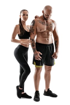 Athletic bald, tattooed man in black shorts and sneakers with beautiful brunette maiden in leggings and top are posing isolated on white background and looking at the camera. Fitness couple, chic muscular bodies, gym concept. The love story.
