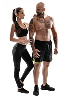 Strong bald, tattooed man in black shorts and sneakers with charming brunette woman in leggings and top are posing isolated on white background and looking at each other. Fitness couple, chic muscular bodies, gym concept. The love story.