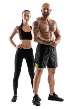 Strong bald, tattooed guy in black shorts and sneakers with charming brunette girl in leggings and top are posing isolated on white background and looking at the camera. Fitness couple, chic muscular bodies, gym concept. The love story.