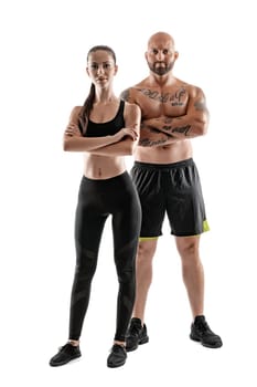 Strong bald, tattooed man in black shorts and sneakers with charming brunette maiden in leggings and top are posing with crossed hands isolated on white background and looking at the camera. Fitness couple, chic muscular bodies, gym concept. The love story.