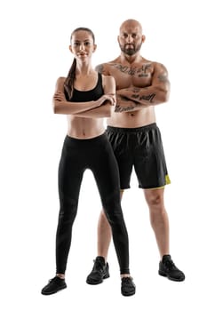 Athletic bald, tattooed man in black shorts and sneakers with gorgeous brunette woman in leggings and top are posing with crossed hands isolated on white background and looking at the camera. Fitness couple, chic muscular bodies, gym concept. The love story.