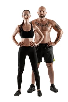 Athletic bald, tattooed guy in black shorts and sneakers with gorgeous brunette girl in leggings and top are posing isolated on white background and looking at the camera. Fitness couple, chic muscular bodies, gym concept. The love story.
