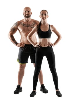 Athletic bald, tattooed fellow in black shorts and sneakers with gorgeous brunette lady in leggings and top are posing isolated on white background and looking at the camera. Fitness couple, chic muscular bodies, gym concept. The love story.