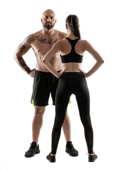 Handsome bald, tattooed man in black shorts and sneakers with attractive brunette woman in leggings and top, standing back, are posing isolated on white background and looking at the camera. Fitness couple, chic muscular bodies, gym concept. The love story.