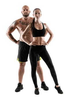 Athletic bald, tattooed man in black shorts and sneakers with gorgeous brunette maiden in leggings and top are posing isolated on white background and looking at the camera. Fitness couple, chic muscular bodies, gym concept. The love story.